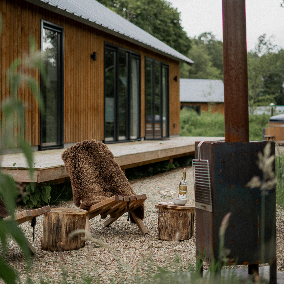 Ein braunes Schafsfell mit Lederschlaufe liegt auf einer Holzbank im Außenbereich vor einem modernen Holzhaus. Daneben stehen rustikale Holzstühle und ein Metallofen, umgeben von grüner Natur, was eine gemütliche, naturnahe Atmosphäre vermittelt.