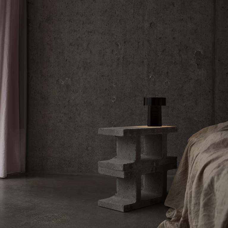 Auf einem modernen, grauen Betontisch steht eine tragbare, schwarze Lampe im minimalistischen Design. Der Raum ist dezent beleuchtet, die Wände sind aus rauem Sichtbeton, ein Bett mit beiger Bettwäsche befindet sich rechts. Tageslicht fällt durch einen zartrosa Vorhang, das Ambiente wirkt ruhig und stilvoll.