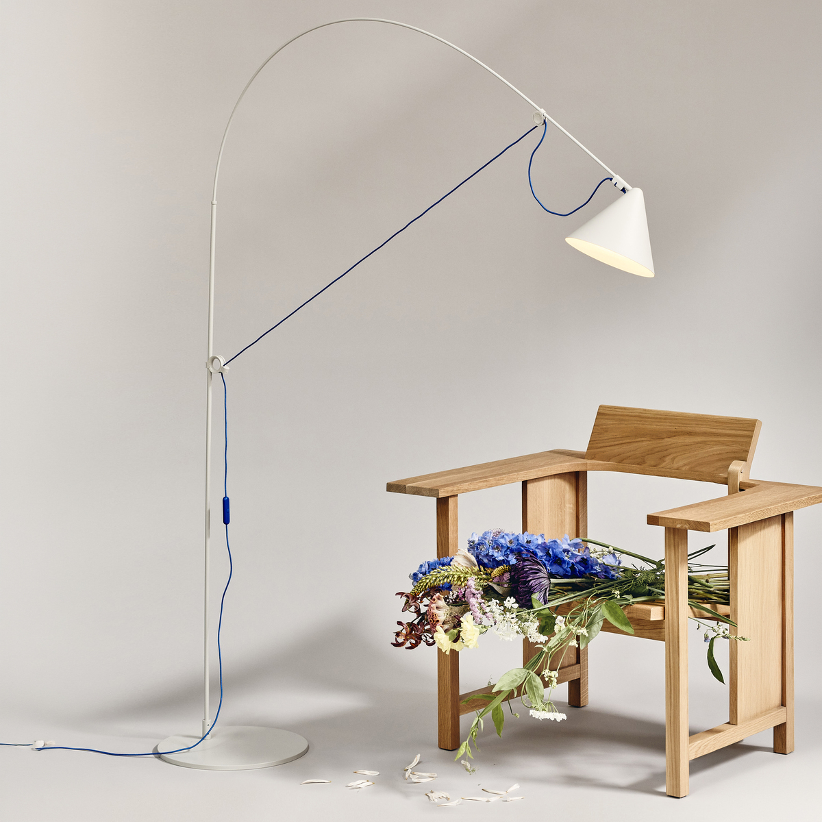 Stehlampe »AYNO« von Midgard in Silk-Grau und Ultramarin mit biegbaren Stab leuchtet stilvoll einen modernen Holzstuhl an, auf dem ein üppiges Blumenarrangement ruht. Die Szene ist minimalistisch und strahlt hochwertige, zeitgenössische Wohnästhetik aus.