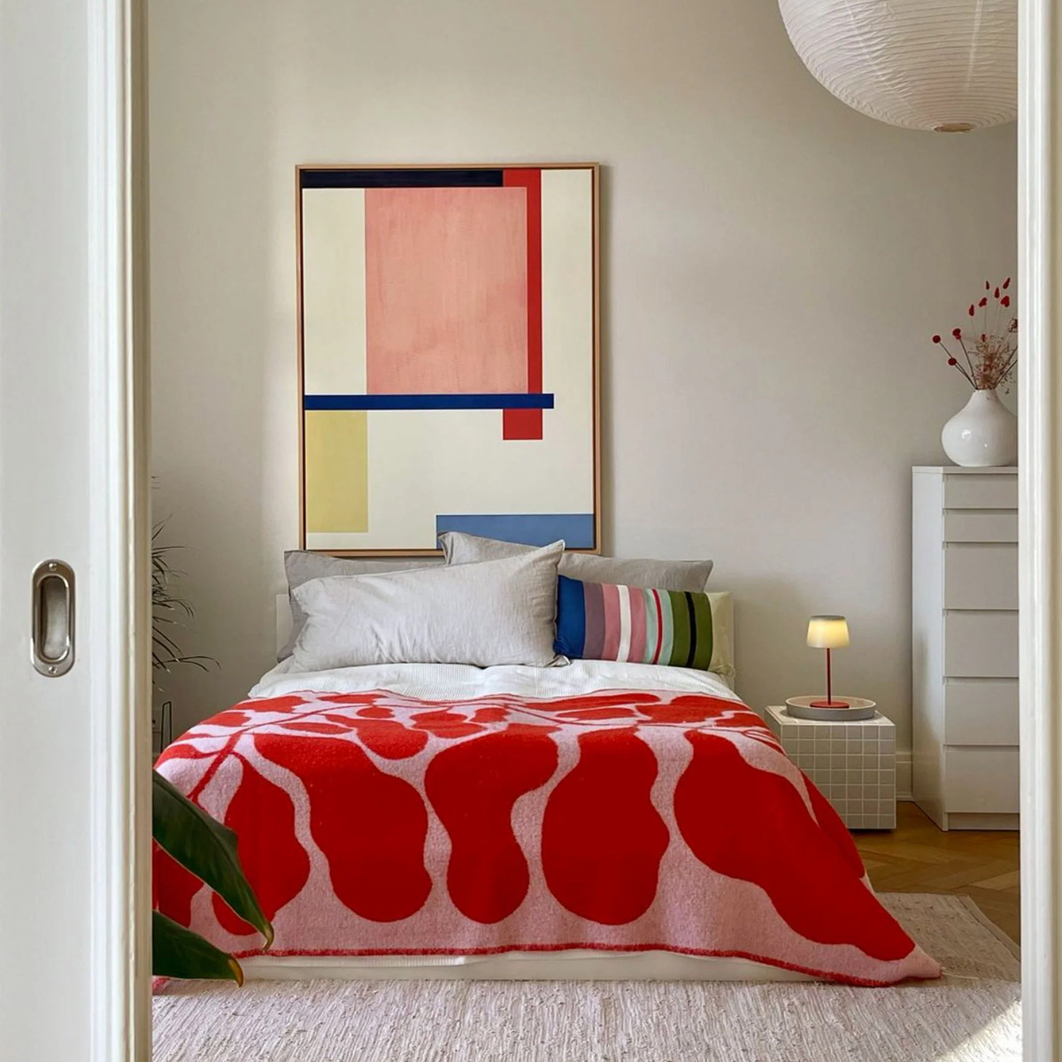 Auf dem Produktfoto ist ein modernes Schlafzimmer mit Holzfußboden zu sehen. Im Vordergrund befindet sich ein Bett mit einer auffälligen, rot-weißen Decke. An der Wand darüber hängt das Leinwandbild »Fokke No 02«, das abstrakte, geometrische Formen in Blau, Rot, Gelb und Rosa zeigt. Der Raum ist minimalistisch eingerichtet, mit einer weißen Kommode, Pflanzen und einer runden Papierlampe.