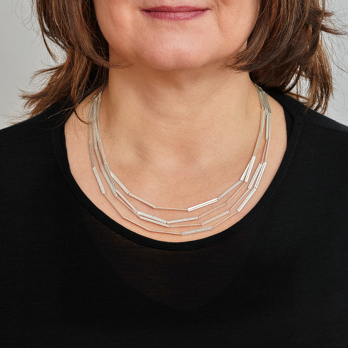 Nahaufnahme einer Frau, die eine silberne Kette mit Streifenmuster trägt. Die Kette liegt auf einem schwarzen Oberteil und besteht aus mehreren dünnen, langen Elementen.