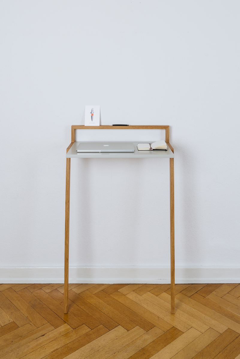 Ein minimalistisches Stehpult »buckowski« aus hellem Holz, das sich ideal für das Homeoffice oder als moderner Arbeitsplatz eignet. Auf der oberen Ablagefläche sind ein kleiner Laptop und ein Notizbuch platziert. Der Pult steht vor einer weißen Wand auf einem Holzboden.