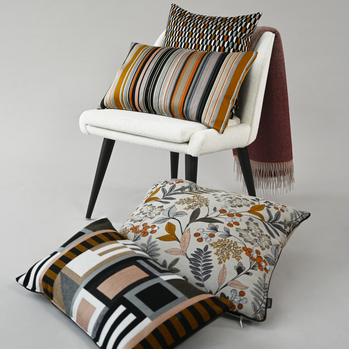 Ein Arrangement von Kissen auf einem weißen Stuhl vor einem neutralen Hintergrund. Die Kissen haben verschiedene Muster, darunter Streifen und florale Designs in Herbstfarben wie Orange, Braun und Grün.