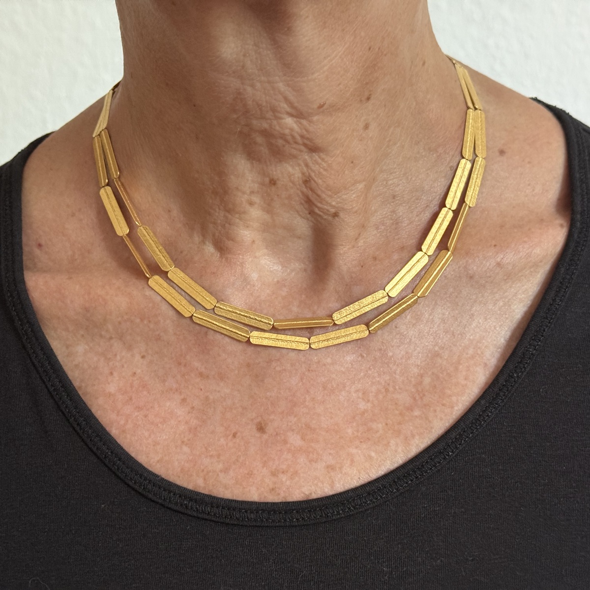 Goldplattiertes Collier aus Silber mit gestreiftem, geprägtem Muster, getragen an einem weiblichen Hals, abgebildet vor neutralem Hintergrund. Das Kettendesign besteht aus mehreren rechteckigen Elementen, die zusammen ein elegantes, modernes Schmuckstück ergeben.