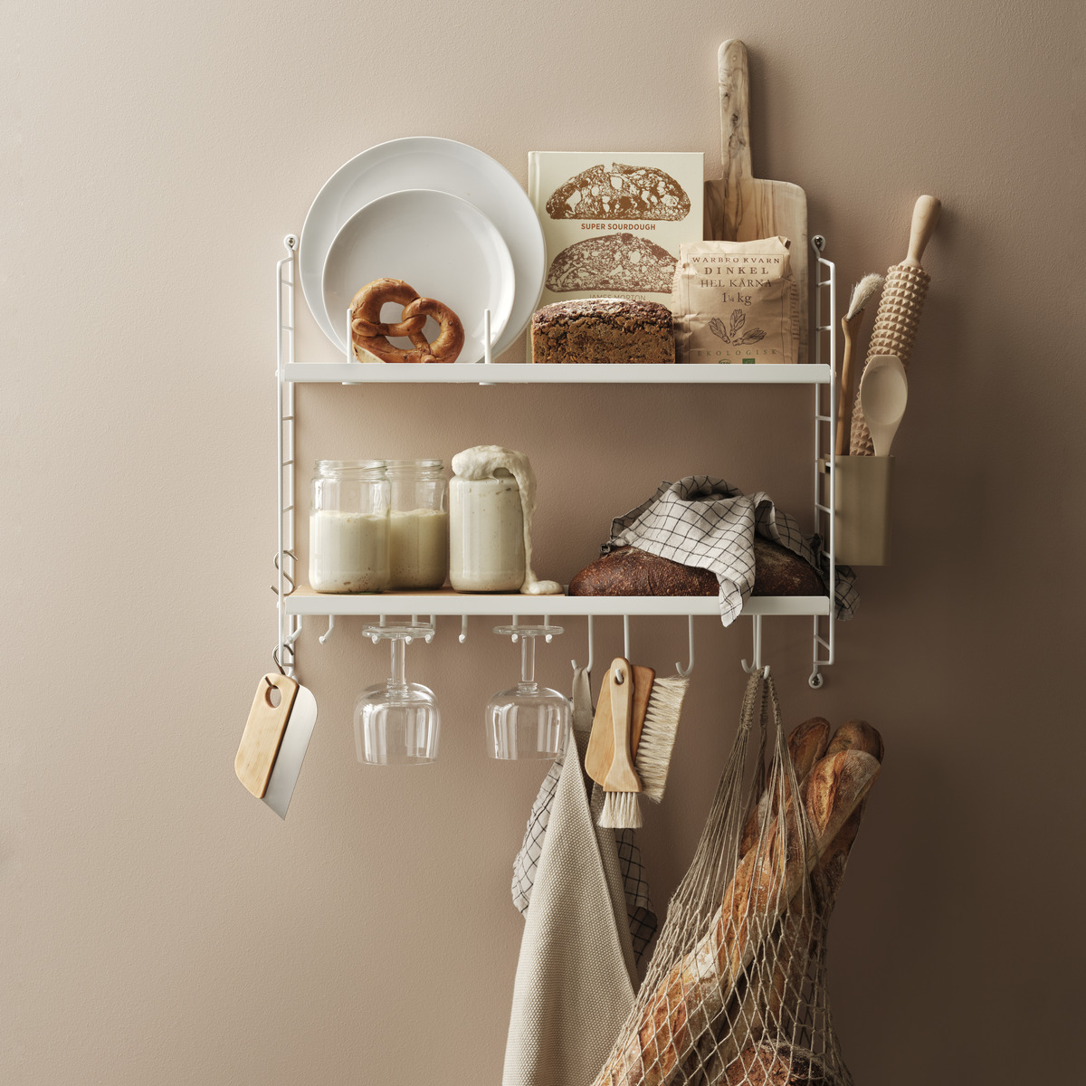 Das Bild zeigt ein weißes, modernes Küchenregal von String an einer beige gestrichenen Wand. Auf dem Regal stehen Schüsseln, Bücher, ein Holzbrett und Gläser. Zudem hängen Geschirrtücher und ein Netz mit Brot am Regal.