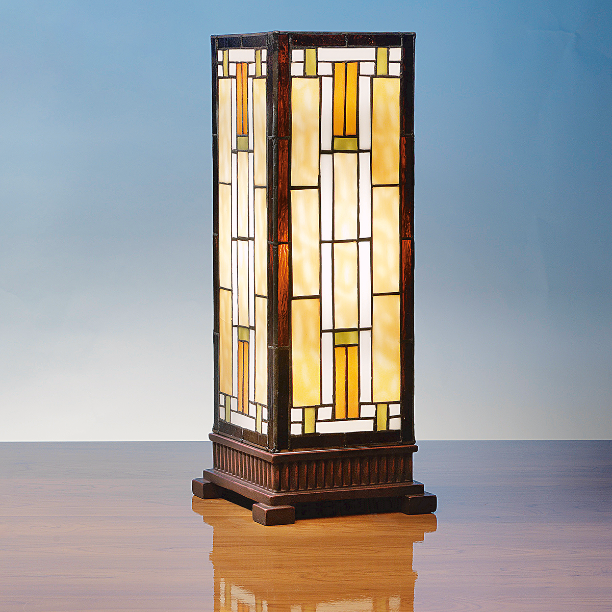 Rechteckige Tischleuchte »Carré« im Stil von Louis C. Tiffany, gefertigt aus mehrfarbigem Tiffany-Art-Glas mit geometrischem Muster in Bernstein-, Creme- und Brauntönen, auf einem kunstvoll verzierten dunklen Kunstguss-Sockel. Die Leuchte steht auf einer glänzenden Holzoberfläche vor blauem Hintergrund.