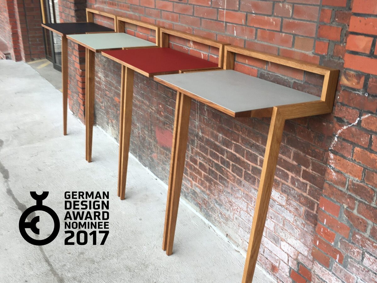 Stehpult 'buckowski' mit modernem Design. Das Pult besteht aus Holz und hat eine Oberfläche in unterschiedlichen Farben: rot, schwarz und grau. Es steht vor einer roten Ziegelwand und hat eine Auszeichnung des German Design Awards 2017 aufgedruckt.
