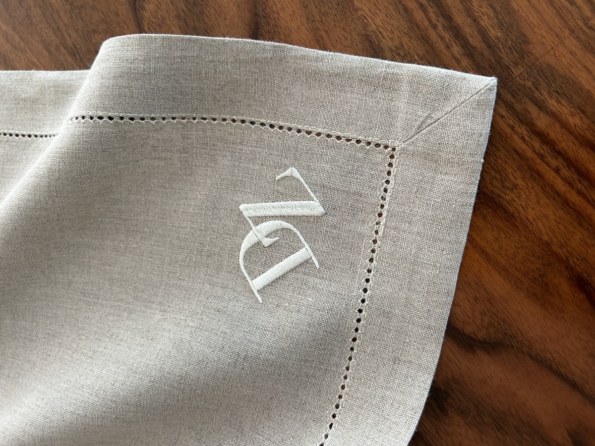 Eine naturfarbene Leinenserviette mit einem aufgestickten weißen Monogramm in der Ecke, auf einem dunklen Holztisch platziert.