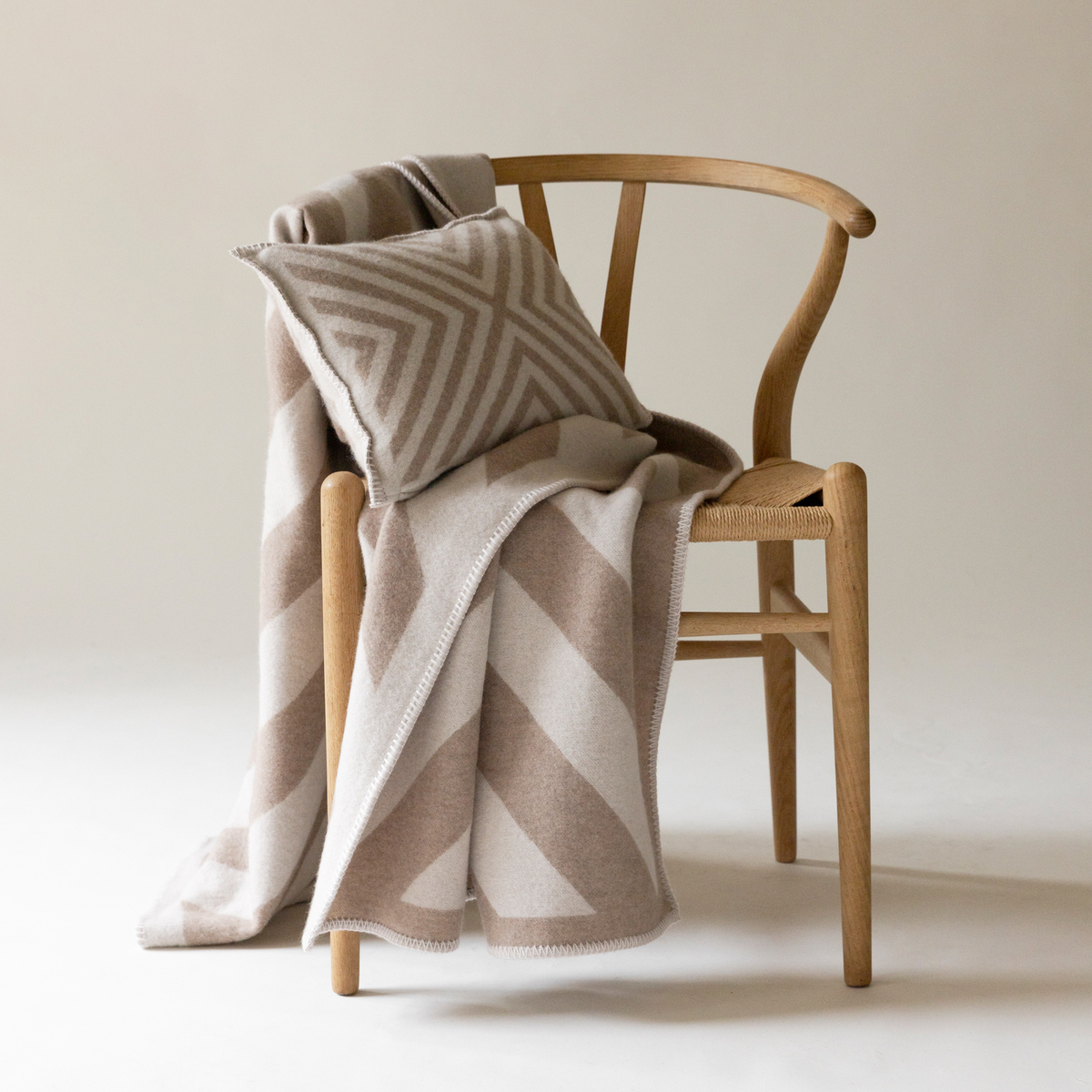 Ein Kaschmir Kissenbezug namens 'Pontresina', platziert auf einem hölzernen Stuhl, kombiniert mit einer weichen Kaschmirdecke in beige und weißen Farben, mit geometrischen Mustern in einem schlichten, eleganten Raum.