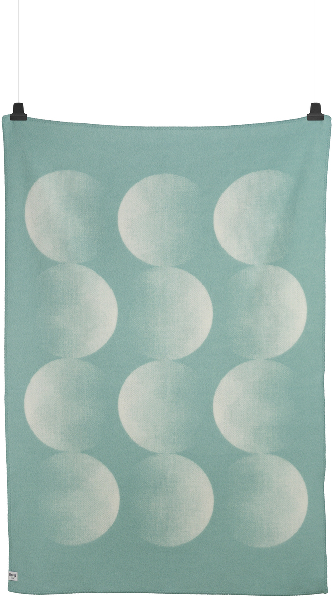 Eine Lammwolldecke namens MOON in Grün mit einem weißen Muster, das an Mondphasen erinnert. Die Decke hängt an zwei schwarzen Clips.