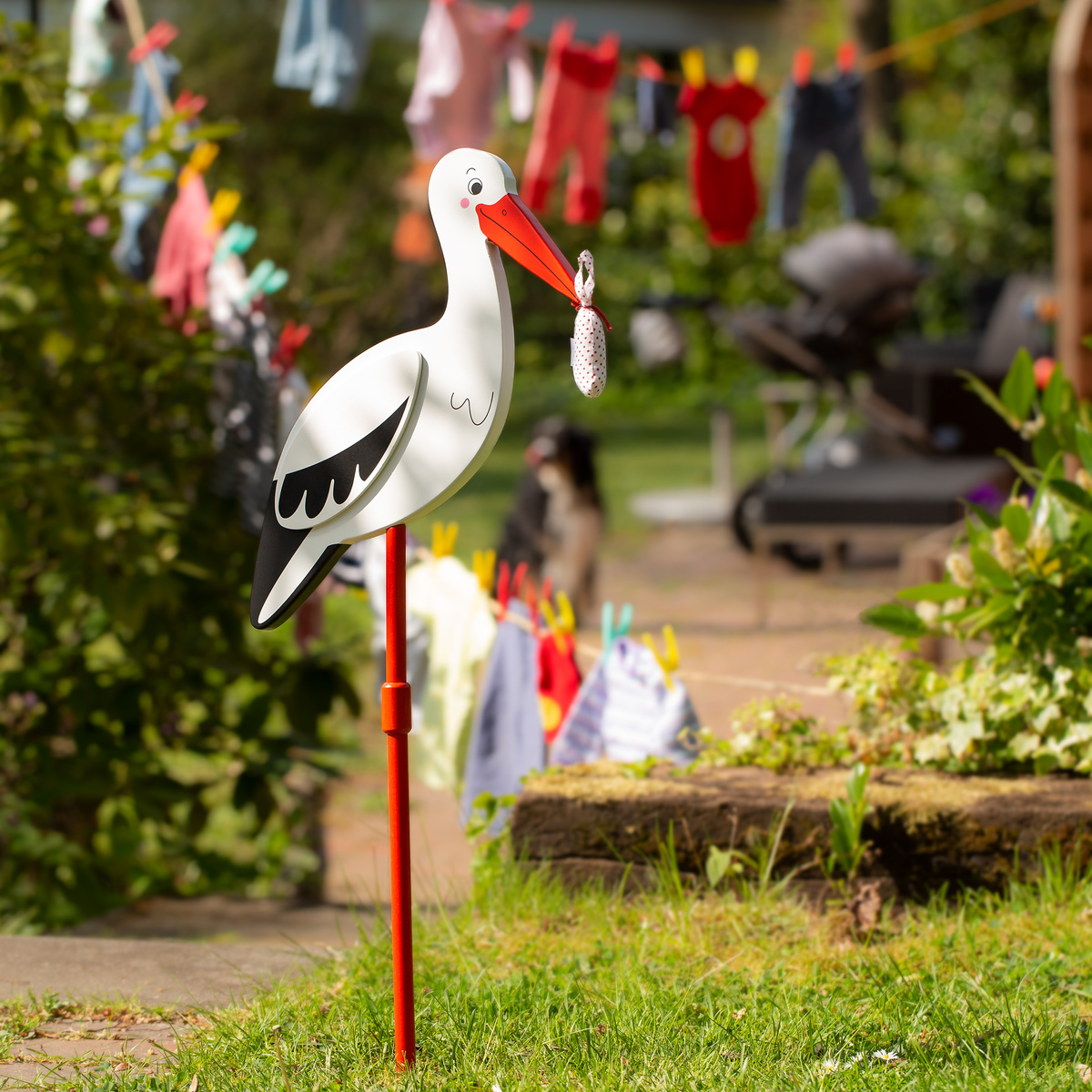 Ein Stecker in Form eines Storchs, der ein Babytuch im Schnabel hält, steht in einem Garten. Im Hintergrund hängt bunte Babykleidung an einer Wäscheleine.