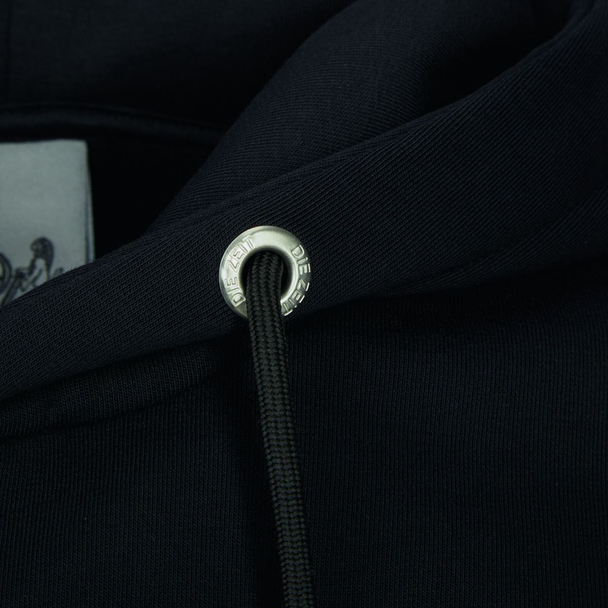 Nahaufnahme eines schwarzen Hoodies aus der ZEIT-Kollektion mit einem edlen Kordelstopper und Detailansicht des Markenlogos.