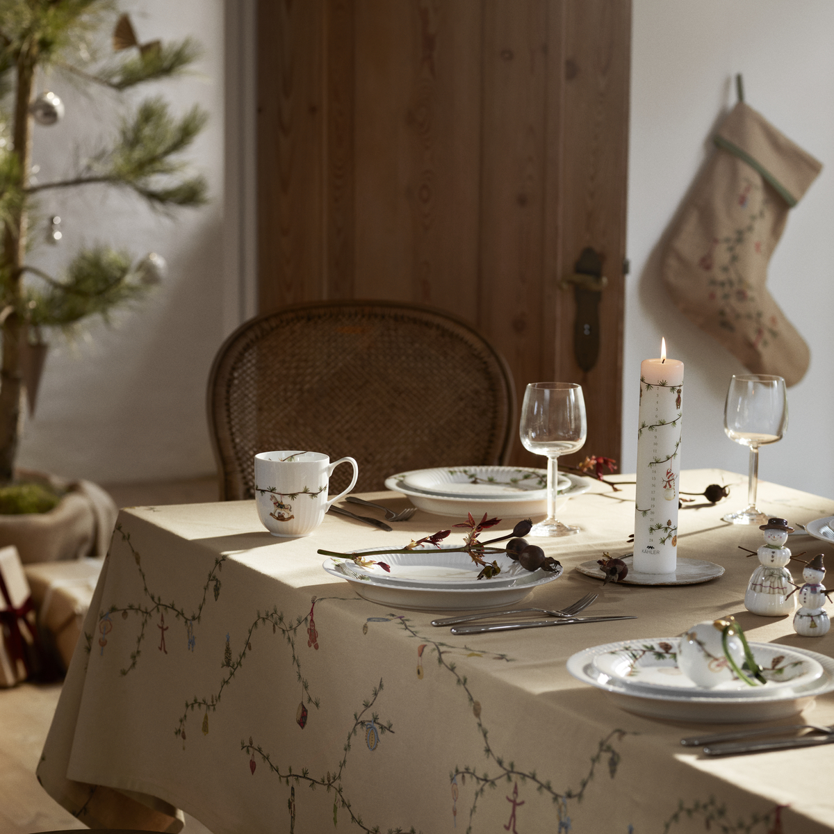 Beige Weihnachtsstrumpf der Marke Hammershøi hängt dekorativ an einer Holzwand in einem weihnachtlich dekorierten Zimmer. Im Vordergrund ist ein festlich gedeckter Tisch mit weiß-beiger Tischdecke und Weihnachtsgeschirr zu sehen. Die gemütliche Atmosphäre wird durch warmes Licht und dezente Weihnachtsdekoration unterstrichen.