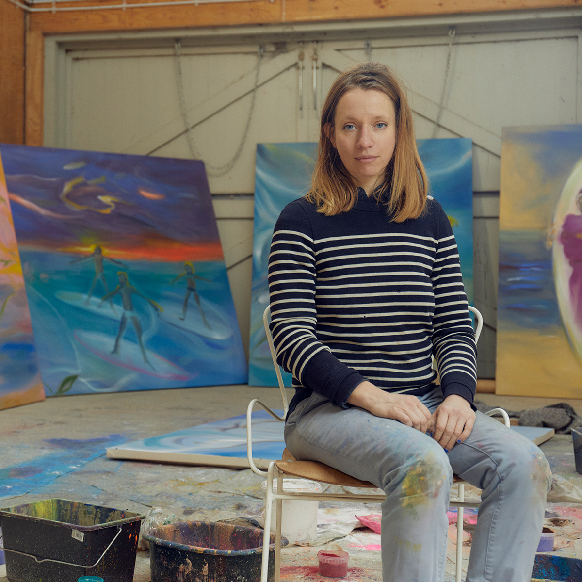 Eine gerahmte Lithografie der ZEIT-Sonderedition »DG 75A« von 2024, präsentiert in einem hell eingerichteten Atelier mit verschiedenen bunten Kunstwerken im Hintergrund. Eine Frau sitzt auf einem Stuhl vor der Kunst und trägt eine gestreifte Pullover.