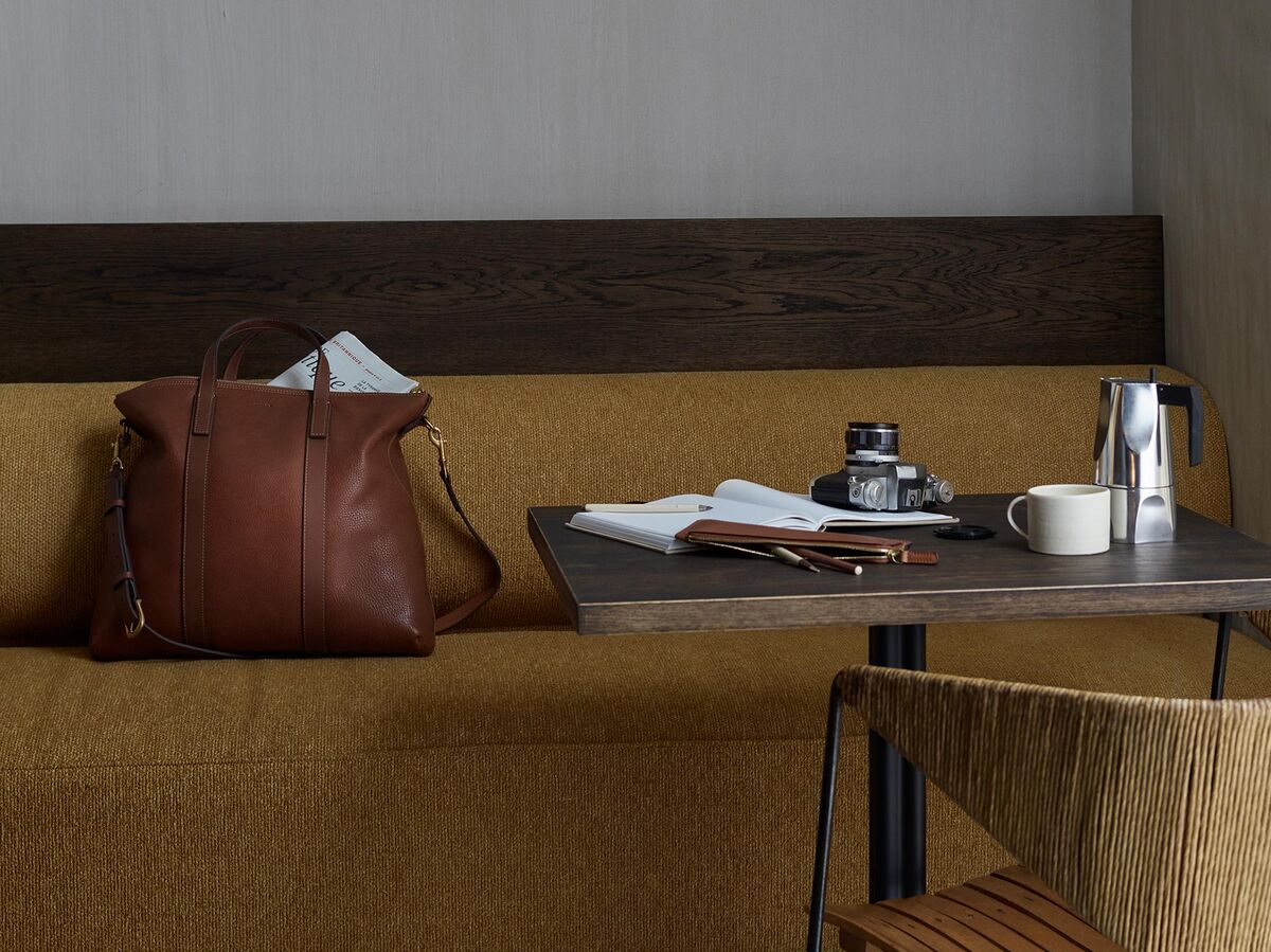 Eine braune Ledertasche »MATE« von Mismo ruht auf einem gelben Sofa neben einem Holztisch. Auf dem Tisch befinden sich Schreibutensilien, ein Hefter, eine Tasse und eine kleine Flasche Tinte. Die Szene hat eine minimalistische und elegante Atmosphäre.