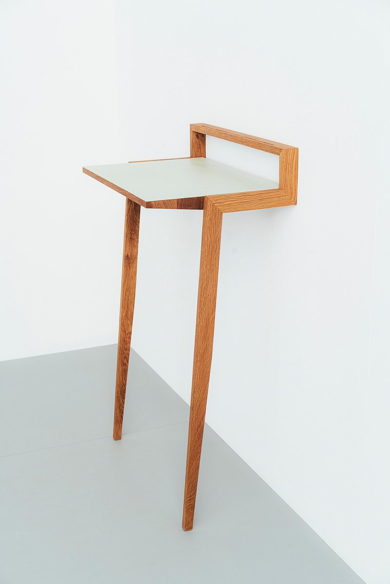 Ein minimalistisches Stehpult aus Holz mit einer weißen Arbeitsfläche. Es hat zwei schmale Beine und ist an eine Wand gelehnt. Der Stil ist modern und platzsparend.