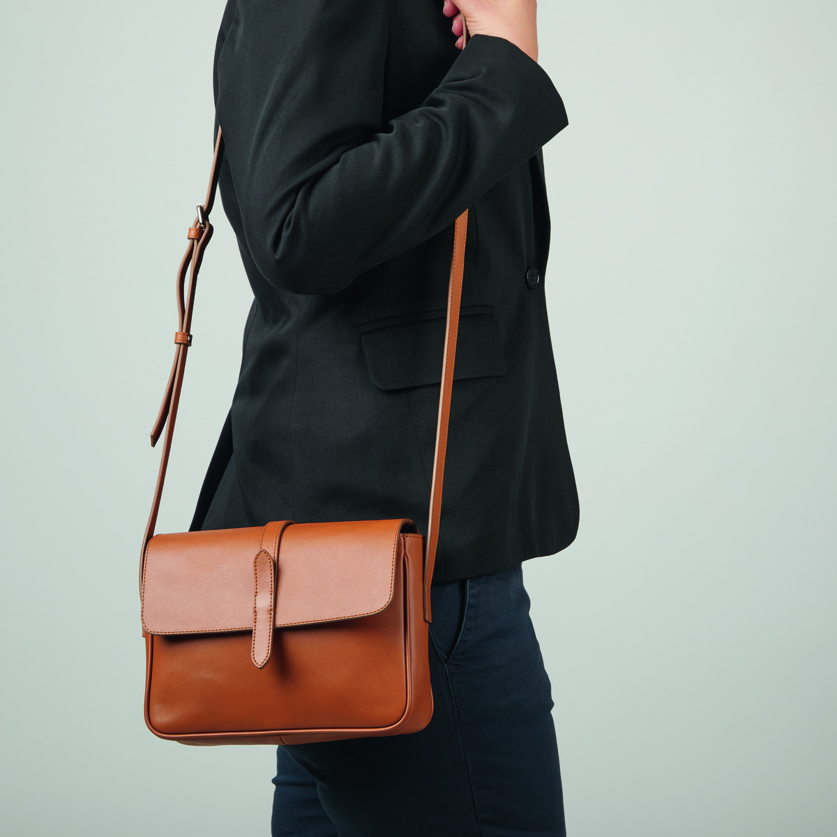 Eine Frau trägt eine ZEIT-Tasche »Saddle Bag Small« in der Farbe Cognac über ihrer rechten Schulter. Die Tasche ist aus Leder und hat ein klassisches, elegantes Design. Die Frau trägt eine schwarze Jacke und eine dunkle Hose.