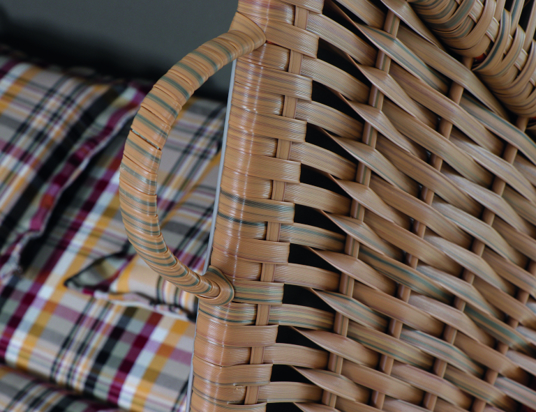 Detailaufnahme eines Gartenstrandkorbs »Lissa« mit geflochtenem Rattan-Material und mehrfarbigem Stoffmuster. Aufgeklappt und sichtbar ist der Griff am oberen Teil des Strandkorbs, daneben liegt ein Kissen mit kariertem Muster in Schwarz, Rot, Gelb und Weiß.