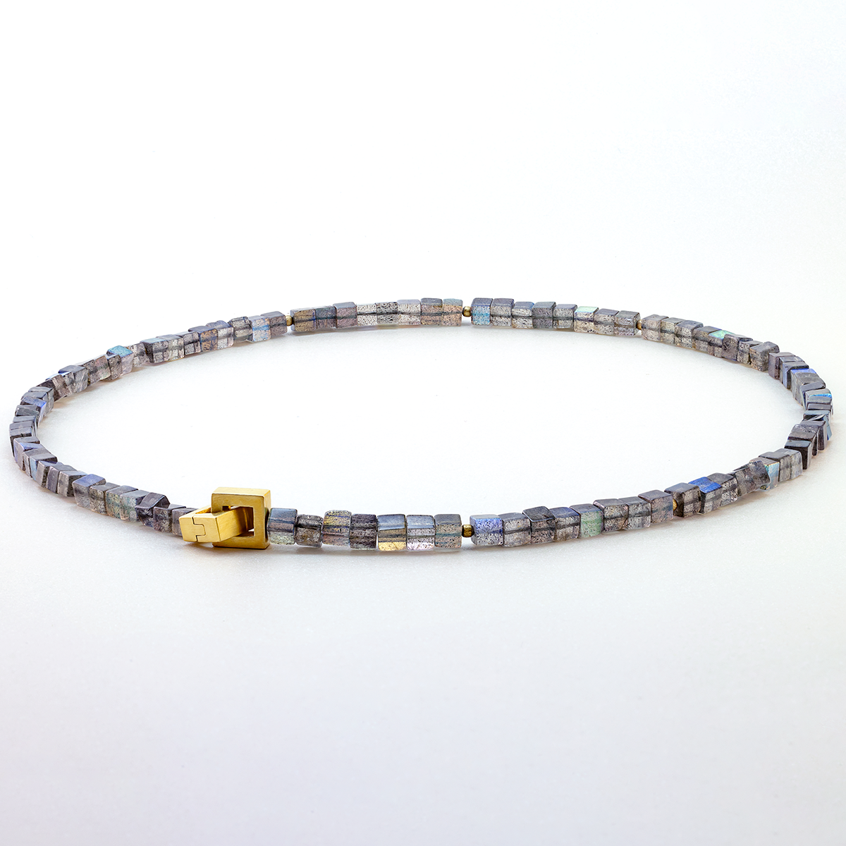 Ein Collier aus Labradorit-Würfeln mit feinem blaugrauem Schimmer, durchsetzt mit mattierten, goldplattierten Silberkugeln. Die Kette besitzt eine dezente, goldfarbene Patentschließe. Das Schmuckstück liegt kreisförmig vor einem weißen Hintergrund.
