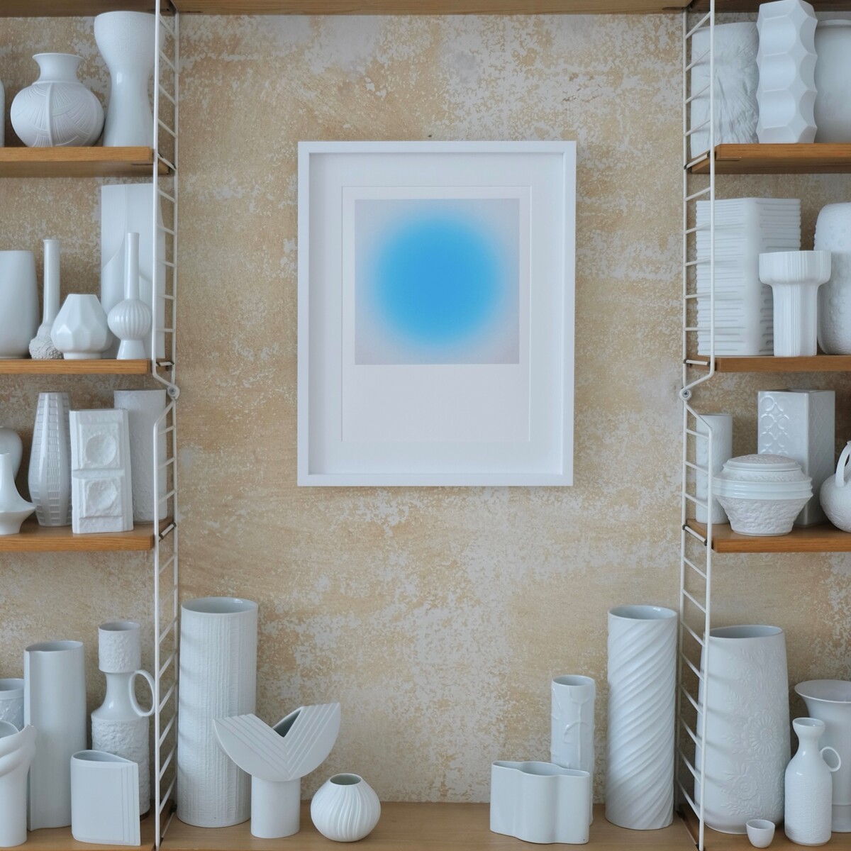 Im Zentrum des Bildes hängt der ZEIT-Sonderedition »Sorbet« Fine-Art-Print von Studio Anastasia Benko an einer beigen Wand mit dezenter Struktur. Das Kunstwerk zeigt einen sanften, blauen Farbverlauf. Beidseitig flankieren zwei Regale mit vielen weißen Keramikvasen, -krügen und -objekten das Bild, die Regalböden sind holzfarben.