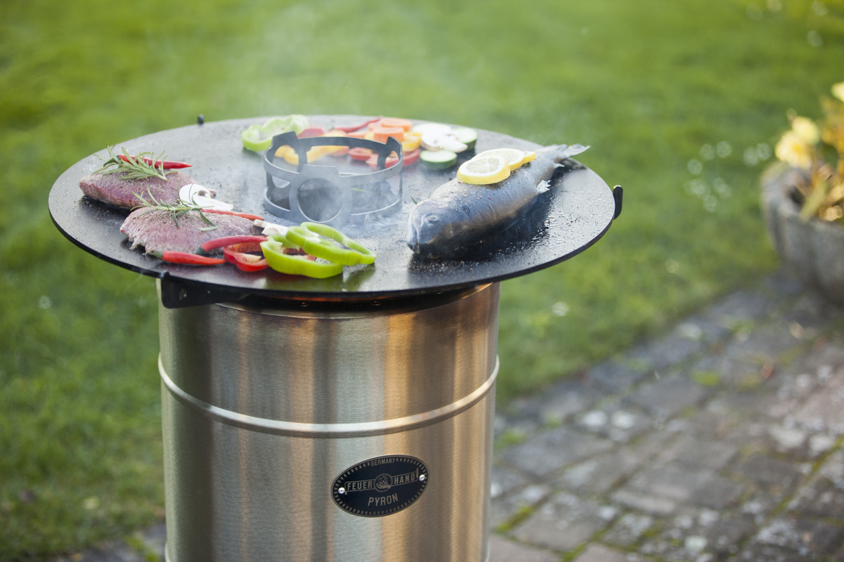Eine Outdoor-Grillplatte von Pyron wird verwendet, um mehrere Lebensmittel gleichzeitig zu grillen, darunter Fisch, Fleisch und Gemüse. Die Grillplatte ist rund und steht auf einem silberfarbenen, zylinderförmigen Sockel. Im Hintergrund befindet sich Grünfläche.