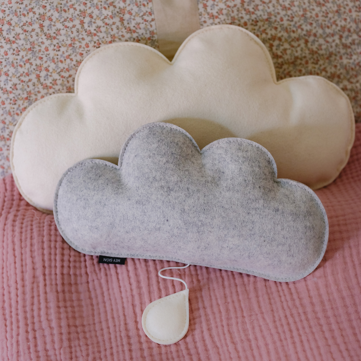 Das Bild zeigt zwei Kissen in Form von Wolken, eines in Beige und eines in Grau, die auf einer rosa Tagesdecke liegen. Im Hintergrund befindet sich ein gemustertes Kissen.