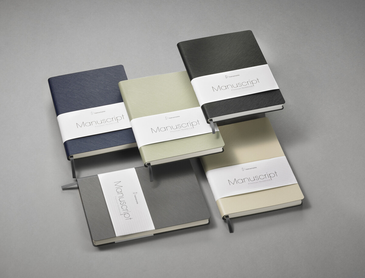 Fünf Notizbücher in verschiedenen Farben (Schwarz, Grau, Beige, Grün) liegen auf einem grauen Hintergrund. Jedes Notizbuch ist mit dem Schriftzug 'Manuscript' und einem schmalen weißen Papierband versehen.