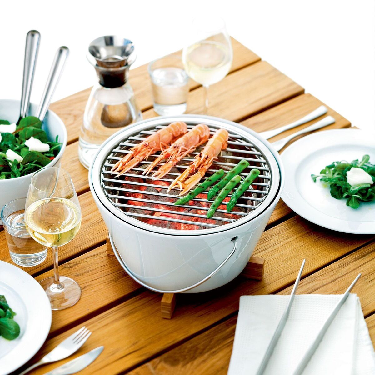 Ein Tischgrill steht auf einem Holztisch, umgeben von Besteck, Tellern mit Salat und Weingläsern. Auf dem Grillrost liegen Garnelen und grüne Bohnen.