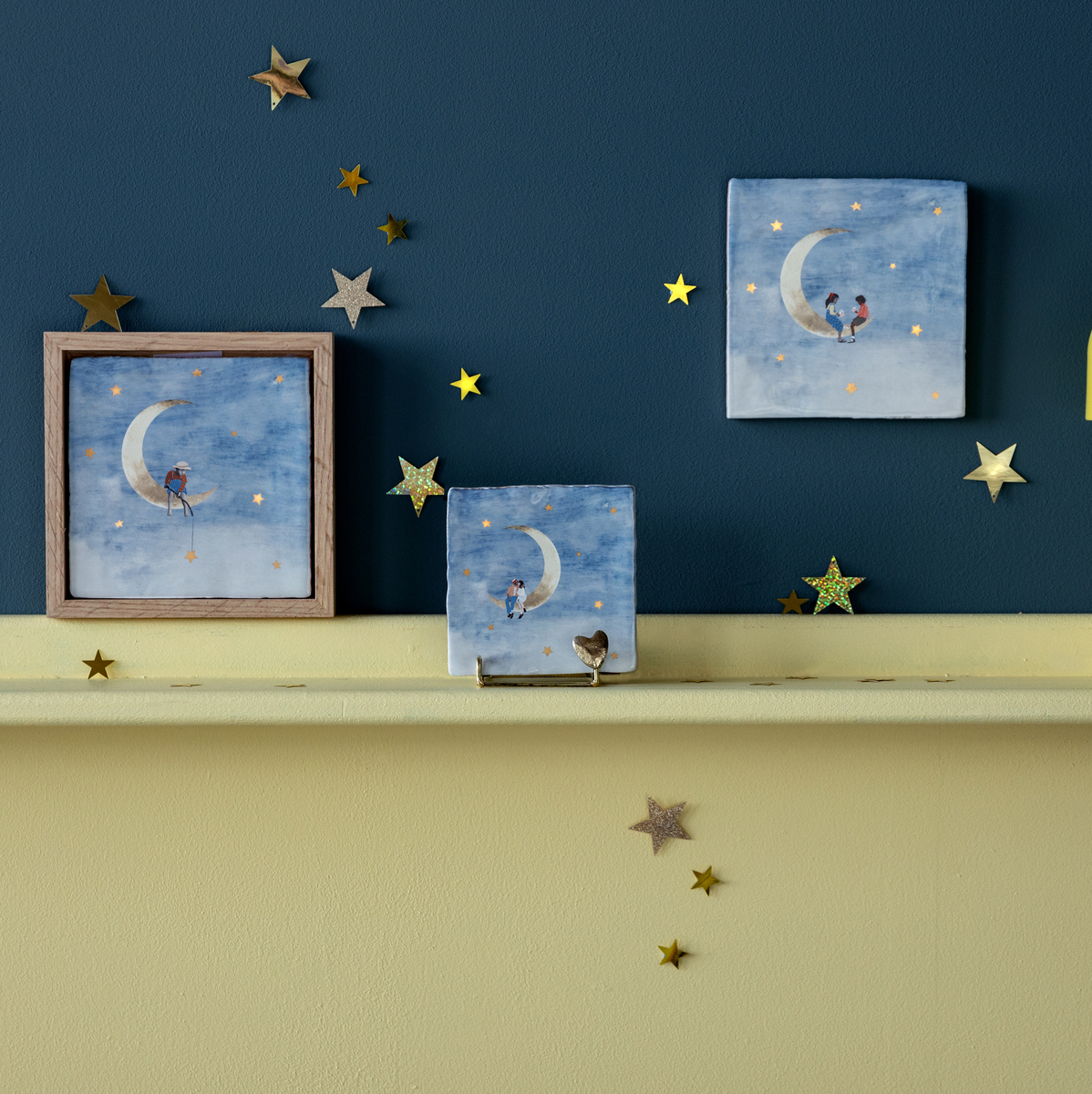 Ein Regal mit einer gelben Lampe und drei Keramikfliesen, die eine Szene von einem Mond mit Sternen und Figuren zeigen. Die Wand dahinter ist blau und das Regal ist gelb. Kleine goldene Sterne sind als Dekoration verteilt.