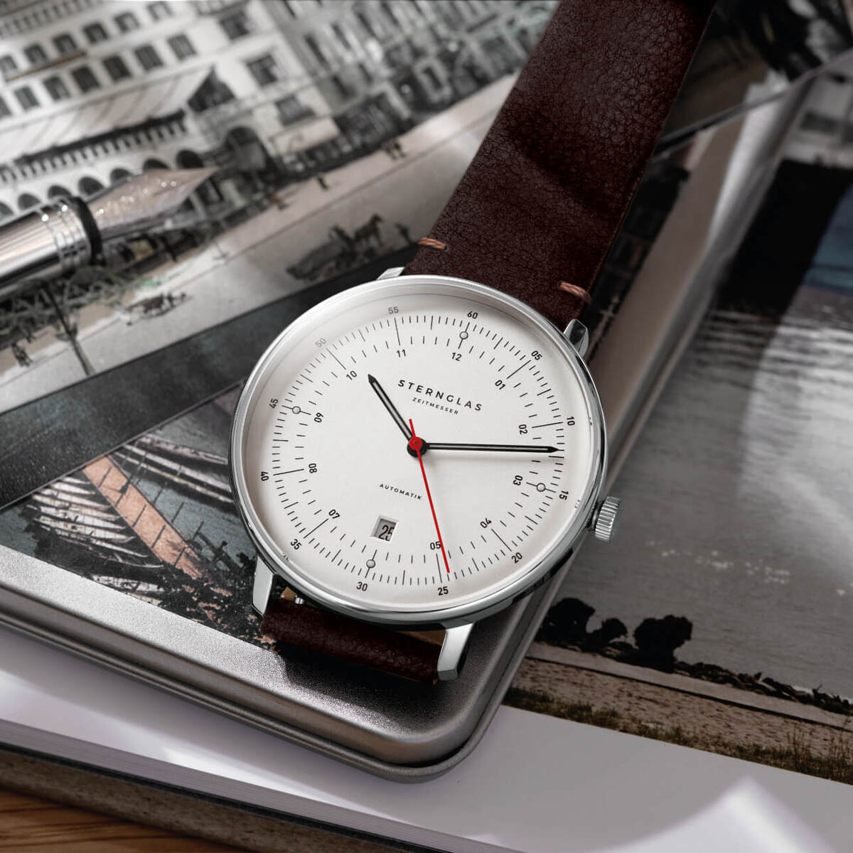 ZEIT-Sonderedition Uhr »Hamburg« mit silbernem Gehäuse und braunem Lederarmband, roter Sekundenzeiger, minimalistisches Design, liegend auf einem Buch und einem Schwarz-Weiß-Foto.