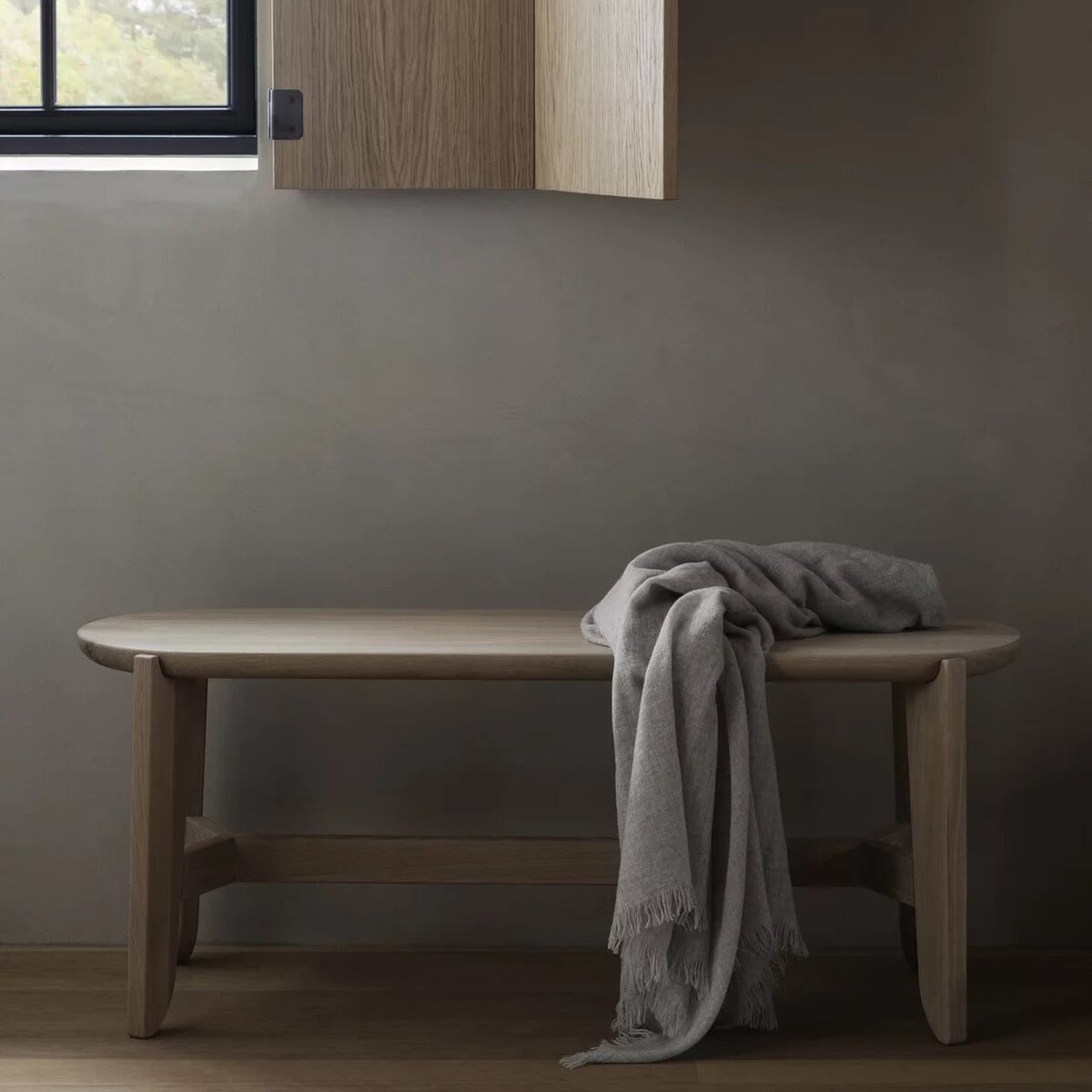 Sitzbank aus hellem Holz im minimalistischen Stil steht an einer Wand unter einem Fenster. Auf der Sitzfläche liegt eine gefaltete, graue Decke. Die Umgebung wirkt modern und ruhig, mit Akzent auf natürlichen Materialien.