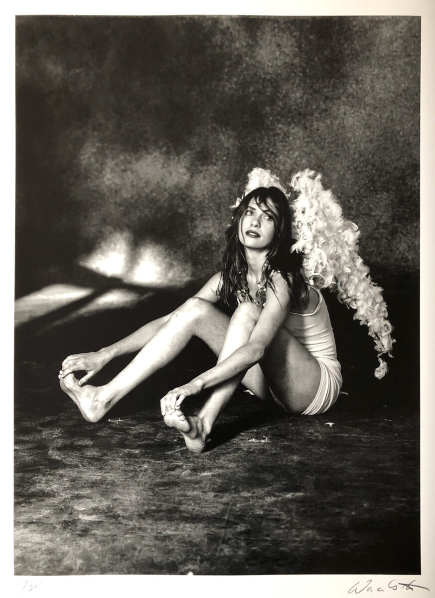 Eine Schwarz-Weiß-Fotografie zeigt eine Frau in sinnlicher Pose mit Engelsflügeln, sitzend auf dem Boden, umgeben von Licht- und Schatteneffekten. Es ist ein künstlerisches Werk mit mystischem Ausdruck, signiert von Wackerbarth.