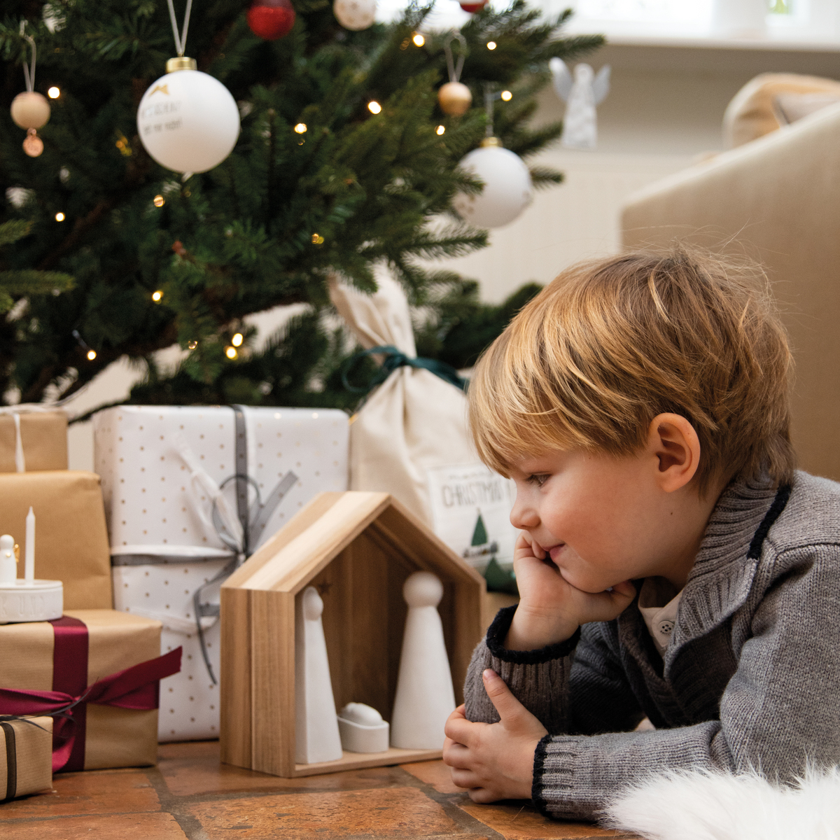 Ein Kind liegt auf dem Boden vor einer Holz-Weihnachtskrippe mit minimalistischen Figuren. Im Hintergrund steht ein geschmückter Weihnachtsbaum mit Lichtern und Kugeln, daneben liegen eingepackte Geschenke.