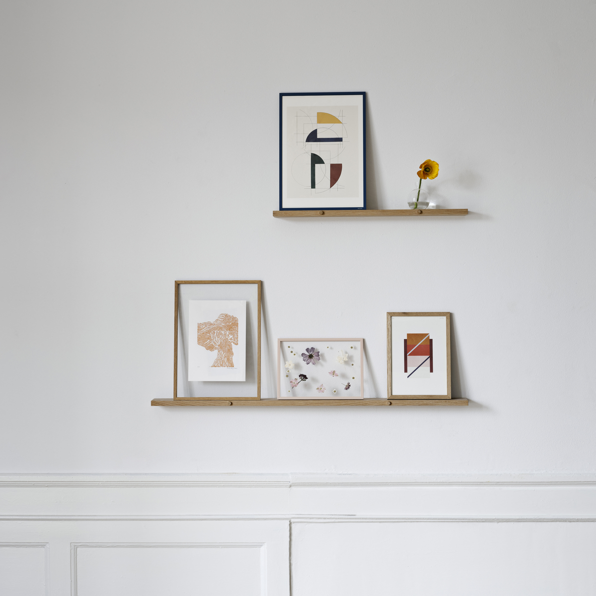 Ein minimalistisches Wandregal aus Eichenholz, das zwei dünne Regalbretter umfasst. Darauf befinden sich mehrere Bilderrahmen mit abstrakter Kunst und eine kleine Sonnenblume in einer Vase. Die Wand ist weiß gestrichen und der Boden besteht aus braunem Parkett.