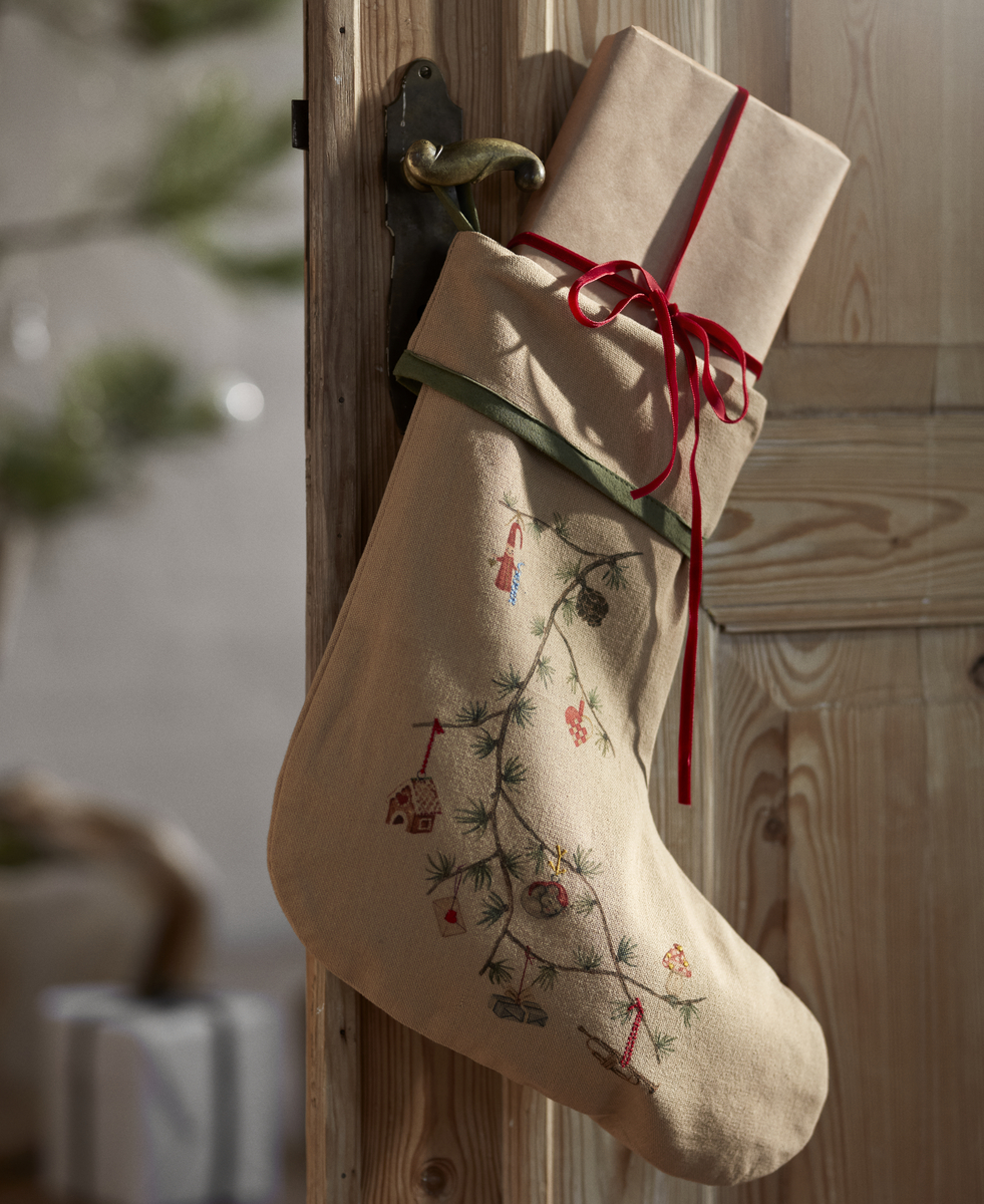 Beiger Weihnachtsstrumpf mit besticktem Tannenzweig und weihnachtlichen Motiven hängt an einer Holztür, darin ein in braunes Geschenkpapier eingepacktes, rotes Band umwickeltes Geschenk. Im Hintergrund ein unscharfer Weihnachtsbaum und ein weiteres Geschenkpaket.