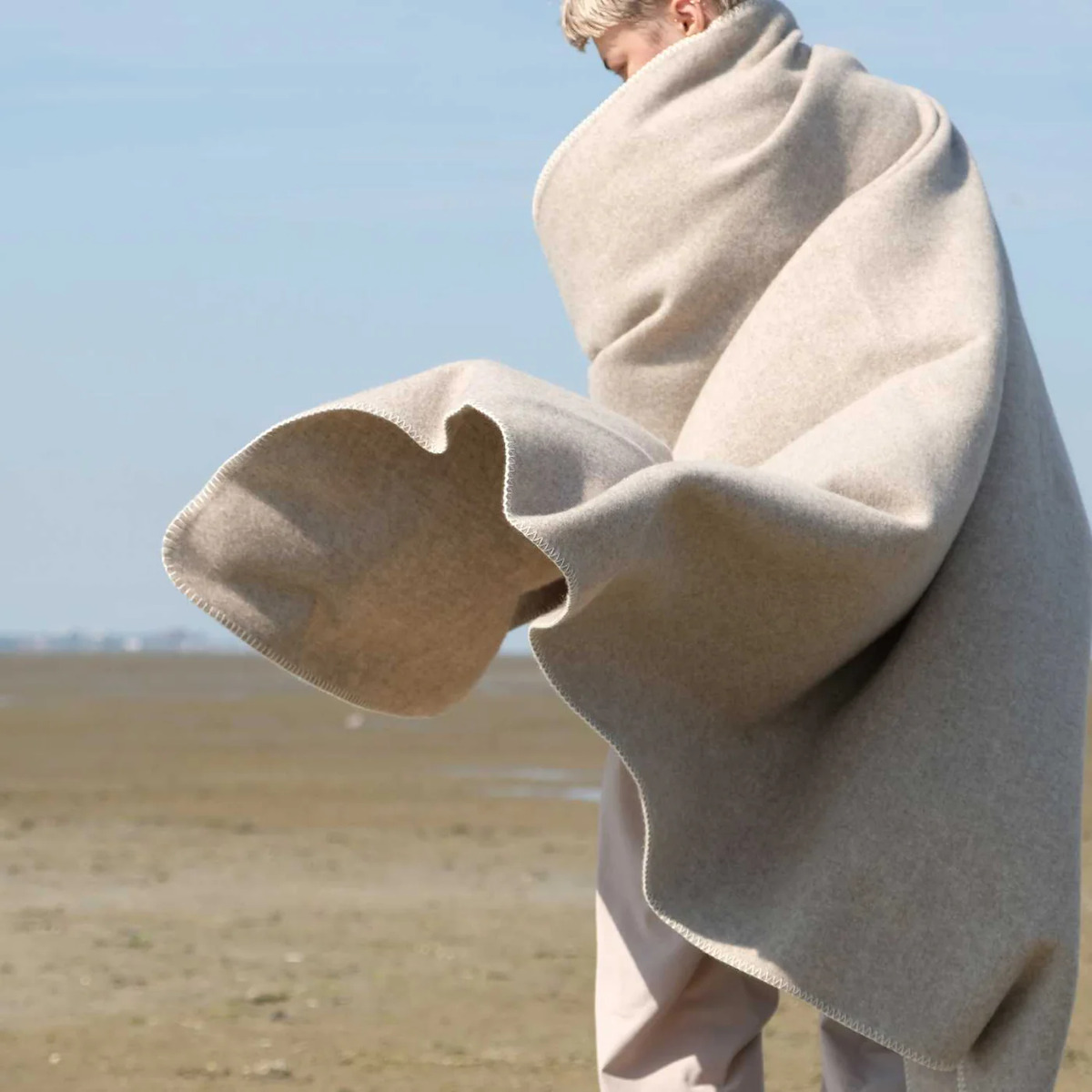 Eine Person ist in eine großzügige hellgraue Merinodecke mit weicher Textur gehüllt und steht auf einer freien, natürlichen Landschaft im Freien. Die Decke hat ein elegantes, minimalistisches Design und vermittelt einen Eindruck von Komfort und Geborgenheit.