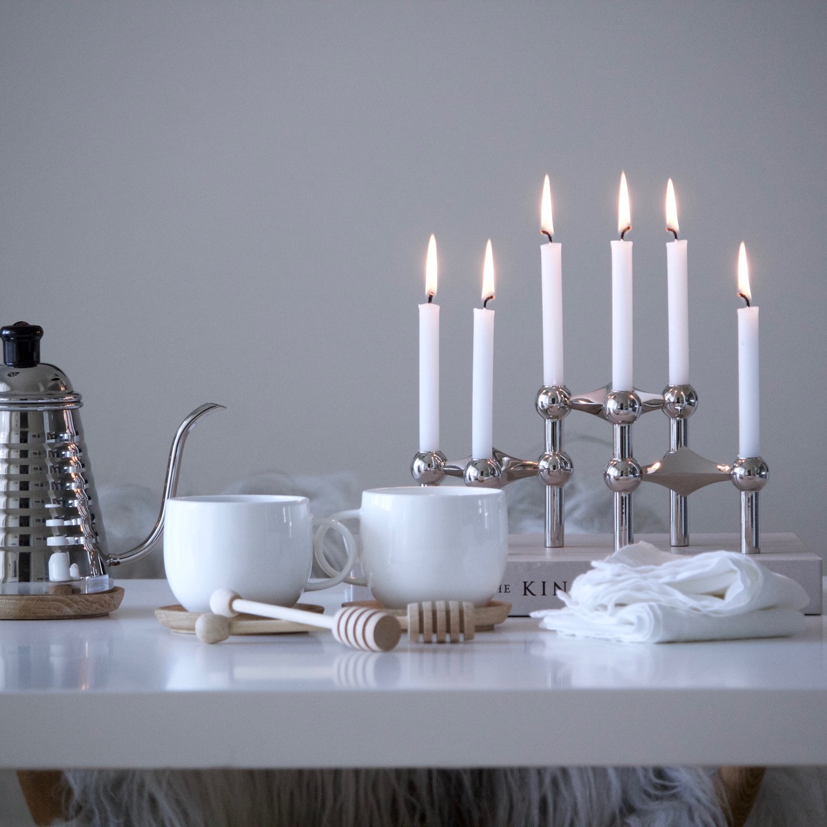 Ein moderner Kerzenständer aus Chrom mit sieben weißen Kerzen auf einem Esstisch, gestaltet mit weißen Tassen, einer Teekanne und Tischdekoration, in einem minimalistisch eingerichteten Raum.