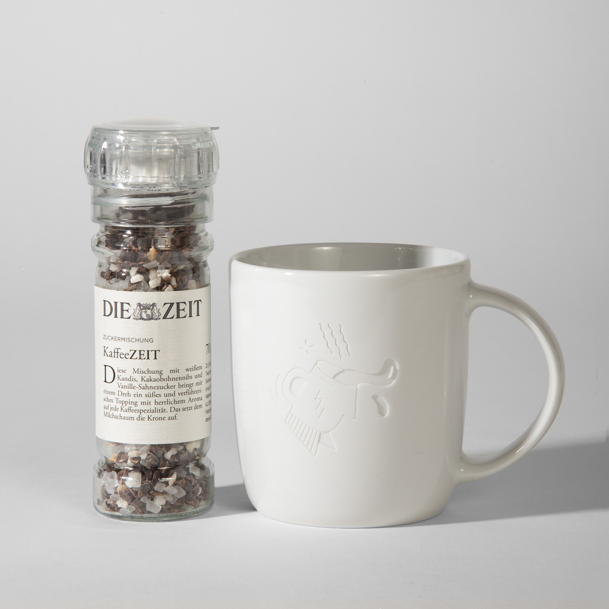 Das Bild zeigt ein Geschenkset, bestehend aus einer Kaffeetasse und einer Gewürzmischung namens KaffeeZEIT von der Marke Die Zeit. Die Tasse ist weiß und schlicht, mit einem dezenten Logo. Die Gewürzmischung ist in einem durchsichtigen Behälter und hat eine schwarze und weiße Farbgebung.