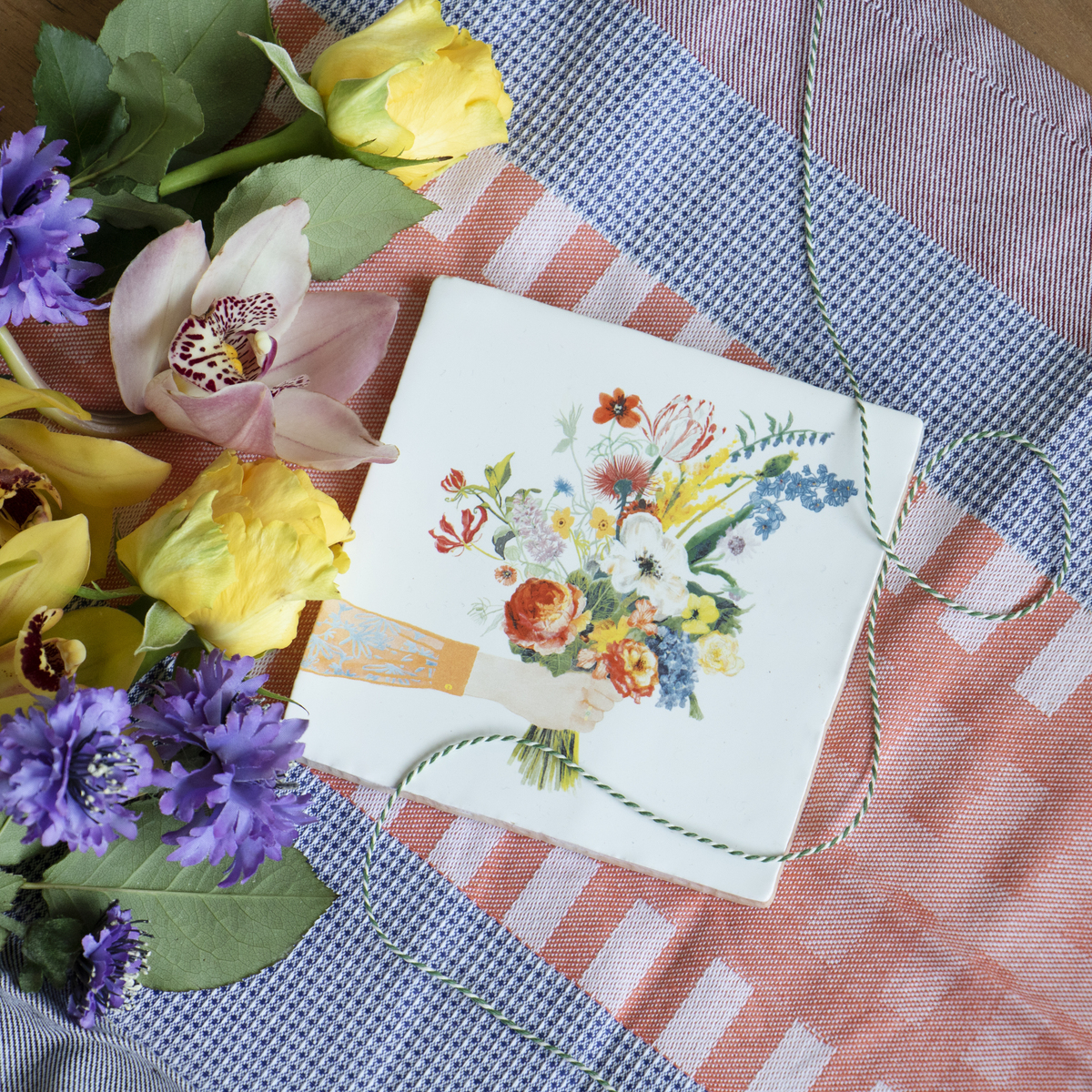 Keramikfliese mit dem Aufdruck eines Blumenstraußes, begleitet von gelben, lila und rosa Blumen auf einem gestreiften Tischtuch.