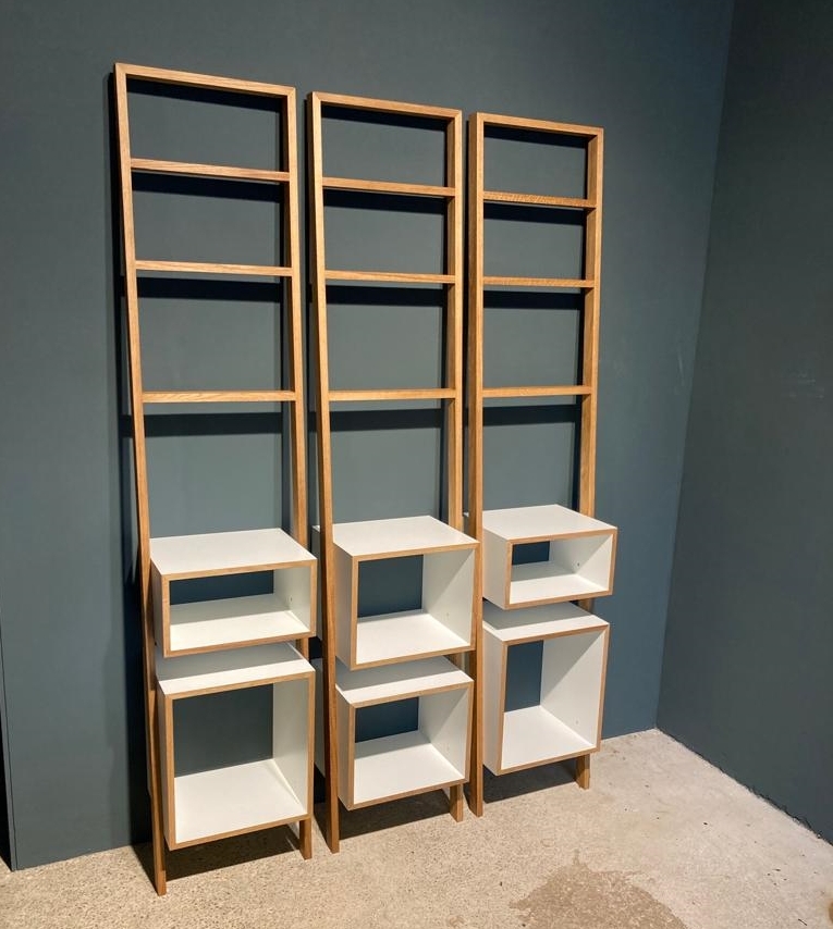 Drei hohe, schlanke Leiterregale »brecht« aus beige-braunem Holz und weißen Regalböden, die an einer dunkelgrauen Wand stehen. Jedes Regal hat mehrere offene Fächer, einige davon quadratisch und andere rechteckig, die flexibel an verschiedene Höhen angepasst werden können.