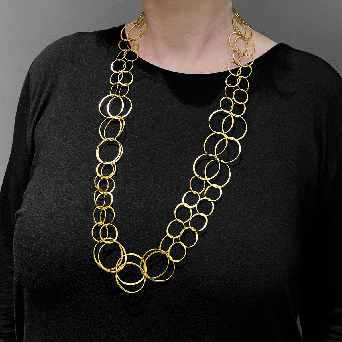 Frau mit blonder Frisur trägt eine lange Halskette mit verbundenen goldenen Ringen aus vergoldetem Silber über einem schwarzen Oberteil. Grau-weißer Hintergrund.