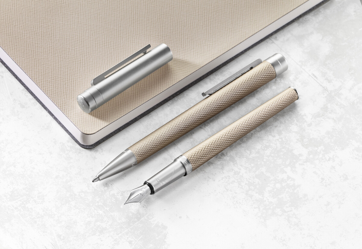 Ein eleganter Kugelschreiber und ein Tintenroller aus der »Slim Edition« liegen auf einer metallischen beigefarbenen Oberfläche neben einem Notizbuch. Beide Stifte sind in silberner Farbe mit einer strukturierten Oberfläche gestaltet.