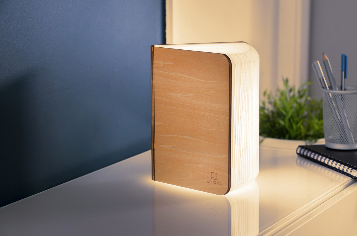 Ein Smart-Booklight in Ahorn-Holzoptik steht auf einem weißen Tisch, umgeben von Schreibutensilien und Pflanzen, und strahlt warmes Licht aus.