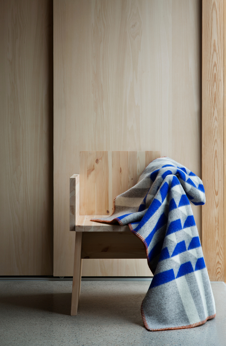 Lammwolldecke »KVAM« in Blau, Grau, Weiß, Beige und Orange mit geometrischem Muster. Drapiert über einem minimalistischen Holzstuhl vor einem hellen Holzhintergrund.