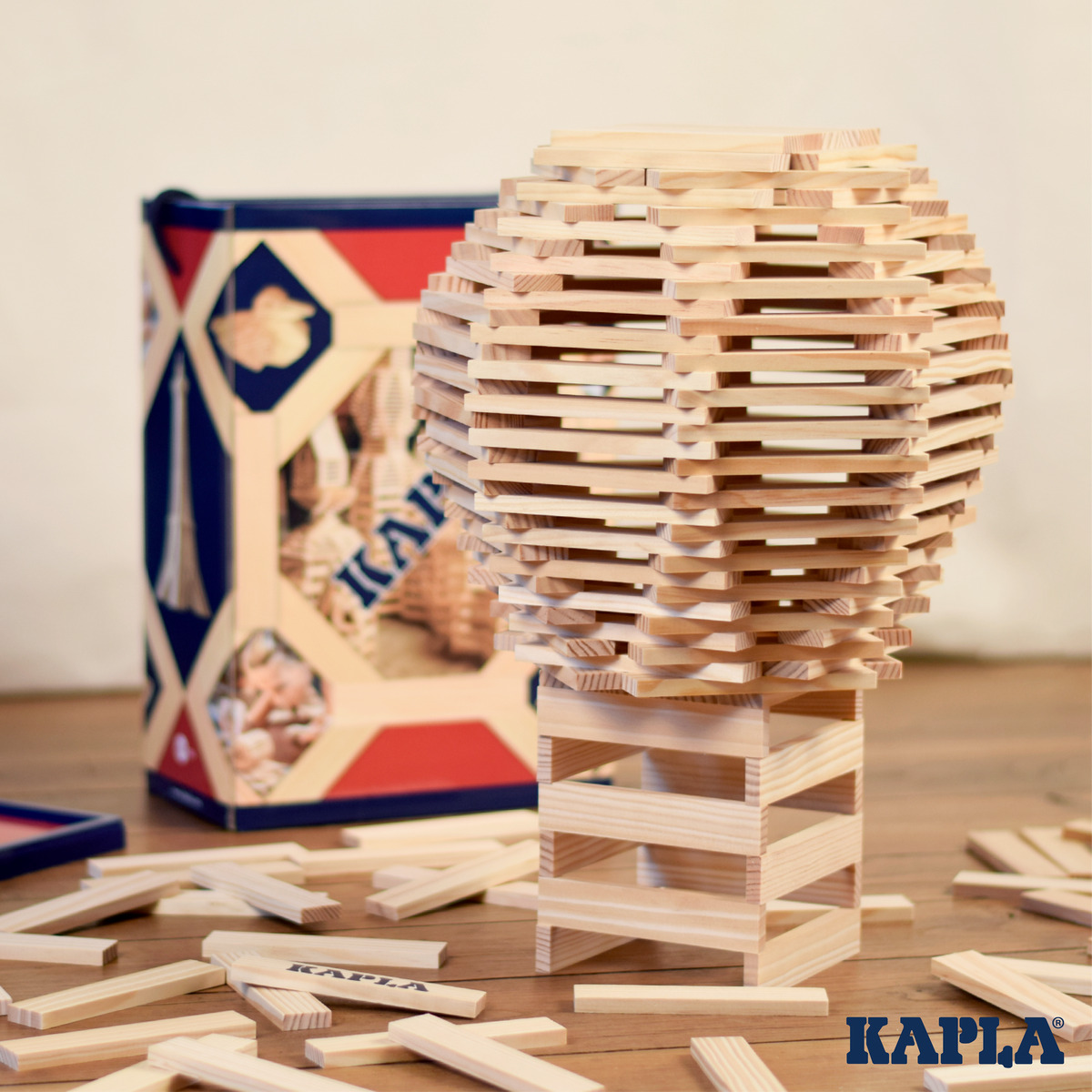 Holzbausteine in einer 200er Schachtel von Kapla. Die Bausteine sind zu einer kreativen Struktur aufgebaut, im Hintergrund ist die Schachtel mit blau-rotem Design zu sehen. Einige Bausteine liegen lose auf dem Tisch.
