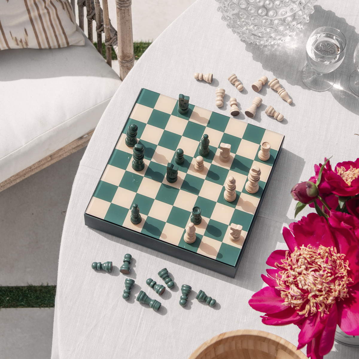 Schachspiel »Classic Chess« auf einem runden Tisch draußen, mit schwarzen und beigen Figuren auf einem beigen und grünen Schachbrett, daneben pinke Blumen und eine Glasdekoration.