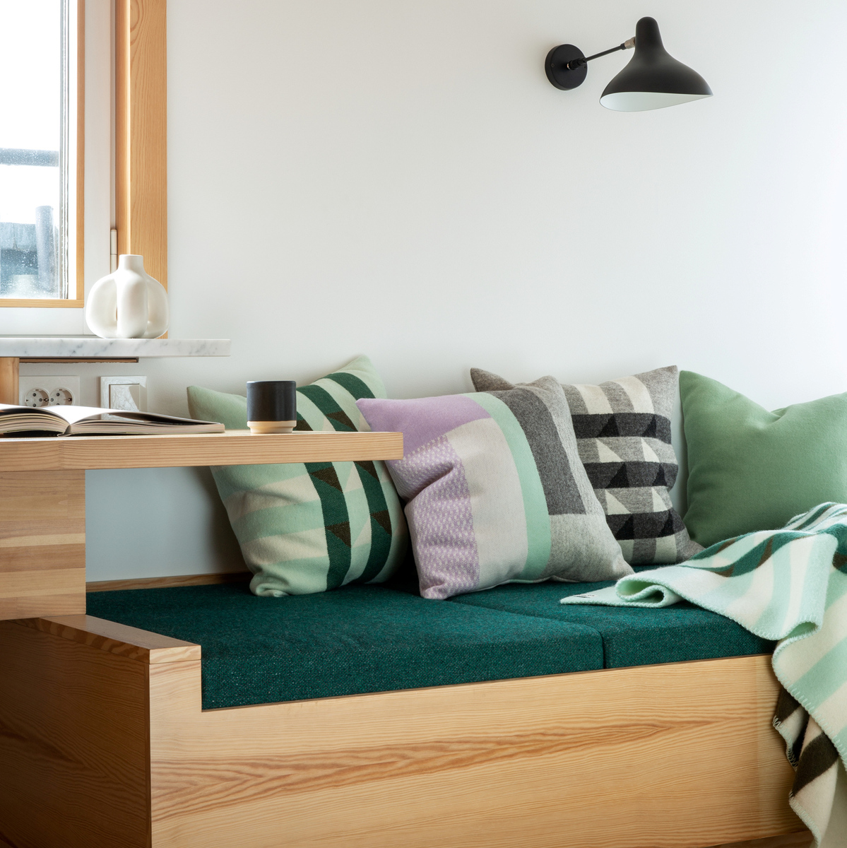 Ein gemütlicher Sitzbereich mit mehreren dekorativen Kissen in verschiedenen Farben und Mustern. Die Sitzbank und der Tisch bestehen aus hellen Holz und daneben befindet sich ein Fenster.