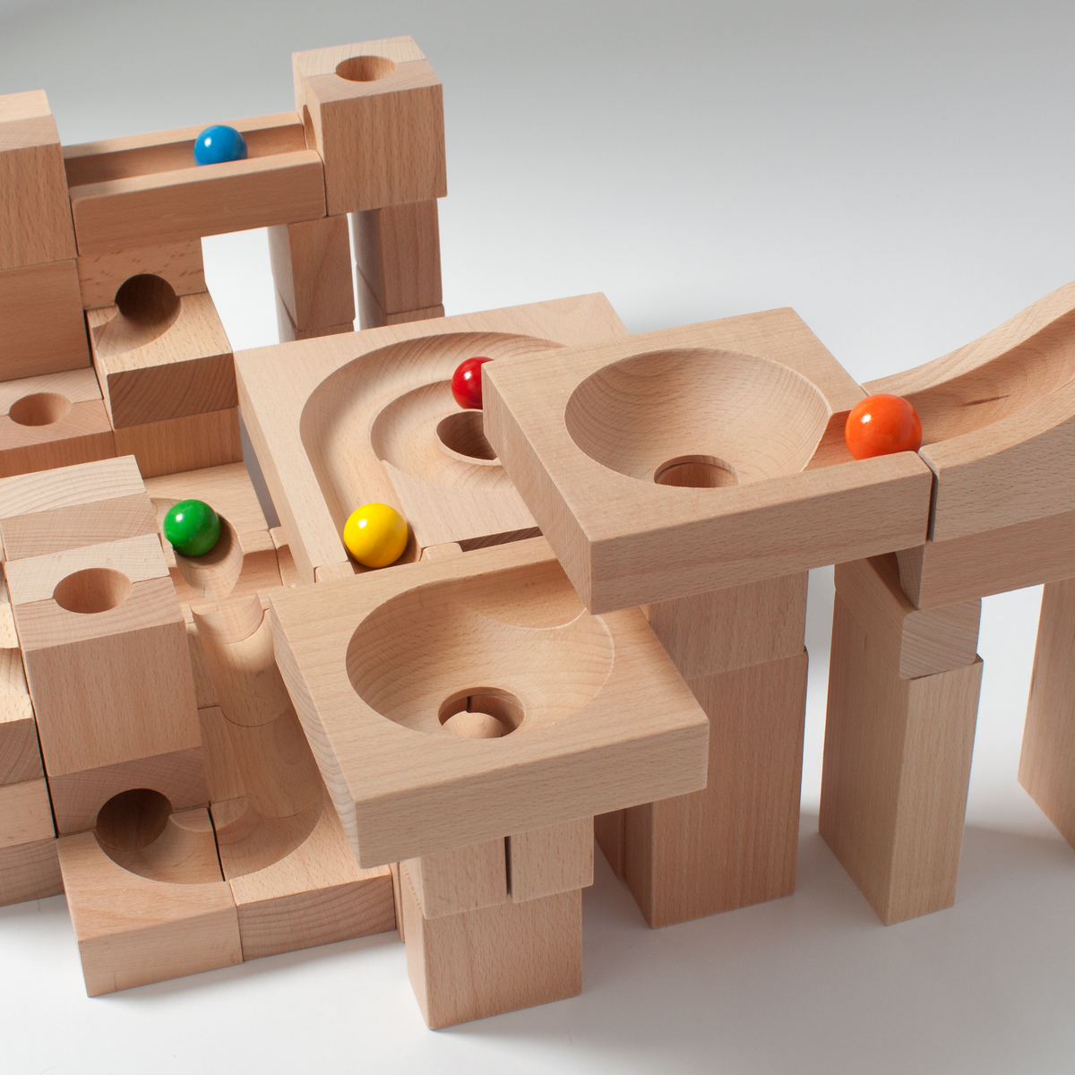 Eine Kugelbahn aus Holz mit bunten Kugeln. Die Konstruktion besteht aus mehreren hölzernen Bauklötzen, die so angeordnet sind, dass die Kugeln durch die Bahn rollen können. Die Kugeln haben die Farben Rot, Blau, Grün, Gelb und Orange.