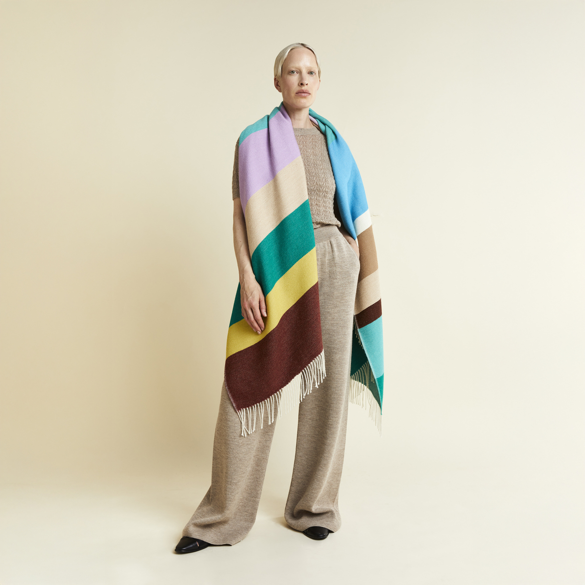 Das Bild zeigt eine Frau mit dem Cape »Mainau« von parati aus 100% Alpaka in Überlänge. Das Cape ist gestreift in Beige, Braun, Türkis, Gelb, Lila und Grün, mit Fransen an den Enden. Die Frau trägt beige Kleidung vor neutralem Hintergrund in elegantem Stil.