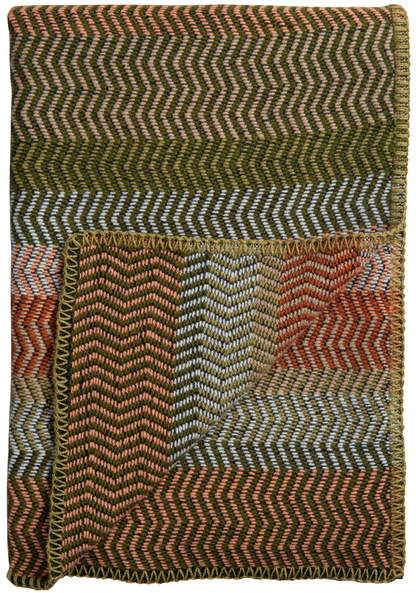 Eine Lammwolldecke mit Zickzack-Muster in verschiedenen Farben wie Grün, Braun, Orange und Weiß. Sie ist typisch nordisch gestaltet, ideal für eine gemütliche Wohnatmosphäre.